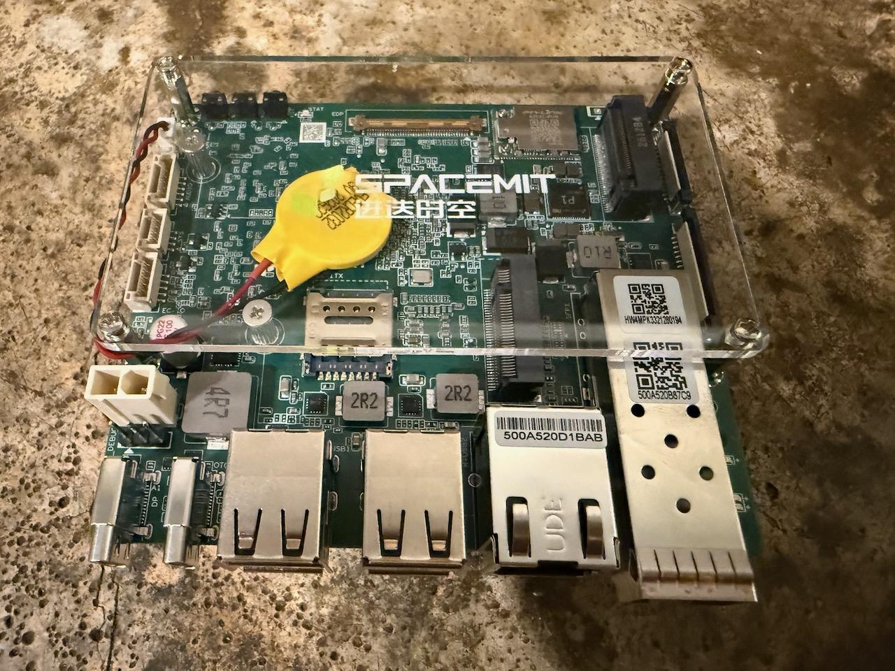 SpacemiT K3 Pico-ITX seen from above under a clear acrylic cover, showing the SpacemiT logo silkscreen, a yellow coin-cell RTC battery, two M.2 slots, a SIM slot and an eDP connector