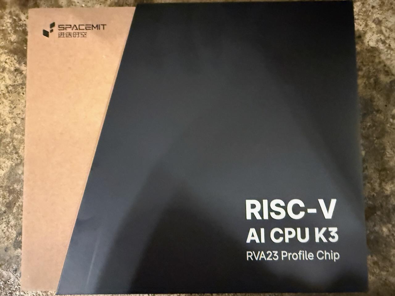 The SpacemiT K3 retail box, black with white lettering reading RISC-V AI CPU K3, RVA23 Profile Chip, with the SpacemiT logo on the kraft sleeve behind it
